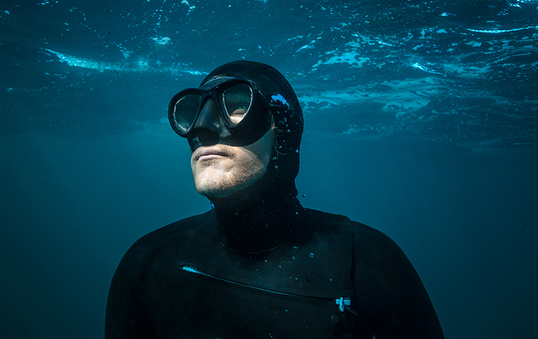 Film still for Astronaut in the Ocean - A freediver wearing a mask and wetsuit floats underwater, looking upward toward the sunlit surface.