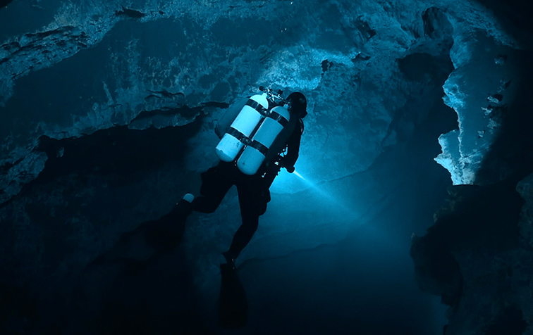 Film still for Diving into the Darkness - A scuba diver with tanks navigates through a dramatic underwater cave or cavern illuminated by blue light.