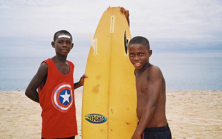 Film still for We The Surfers - Two young boys pose with a yellow surfboard on a beach, one wearing a Captain America tank top.