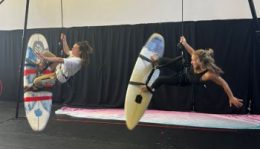 In rehearsal, two dancers are suspended on aerial wires with their feet on surfboards, also suspended
