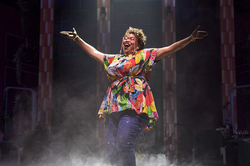 Karen Mavundukure - a black woman with afro hair and a big smile, stands with arms outstretched, singing