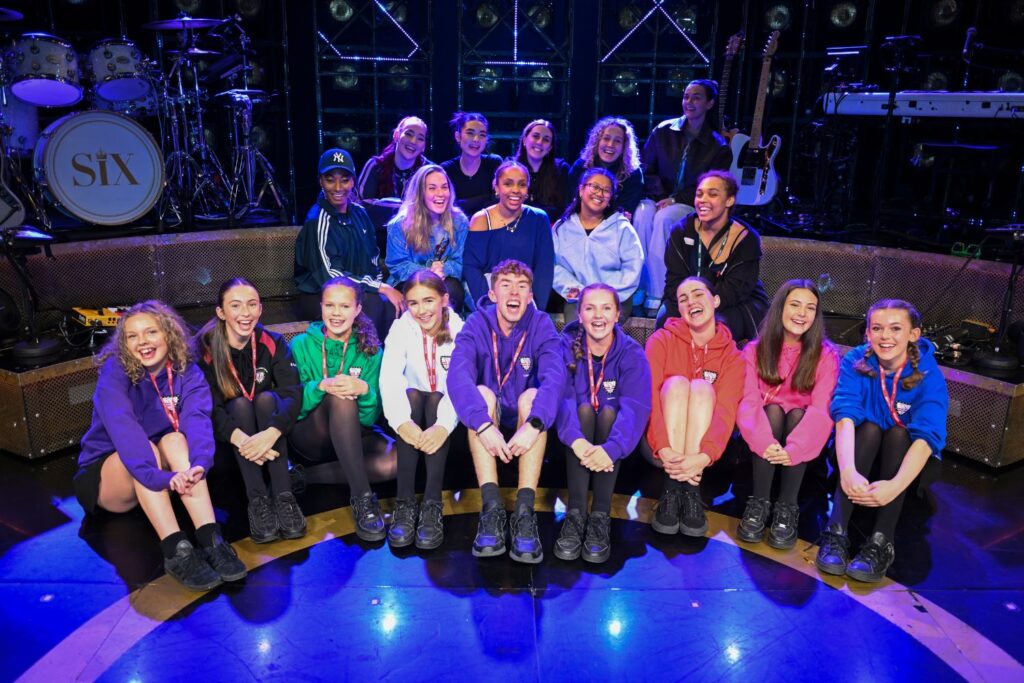 A large group of students from Richard Lander school and professional cast from SIX The Musical sit on the SIX set on the Cornwall Playhouse stage