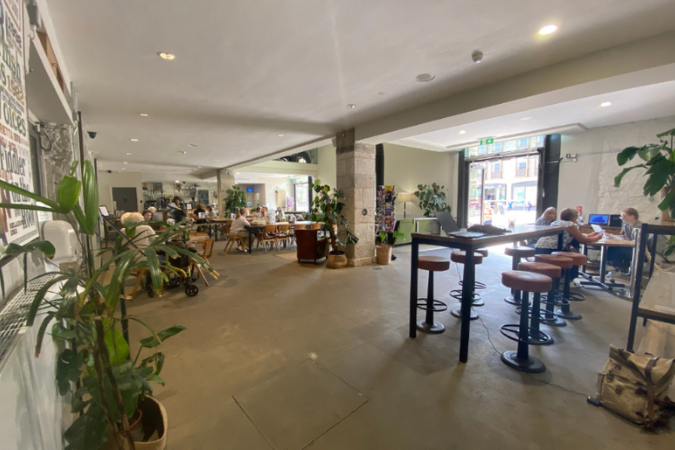 A photo of the Green Room Cafe taken from a corner on a wide lense. A series of tables and chairs are visible, as well as the entrances. Some plants and greenery are scattered about, as well as a few customers sitting at the tables. You can see the Box Office counter in the righthand corner towards the entrance. 