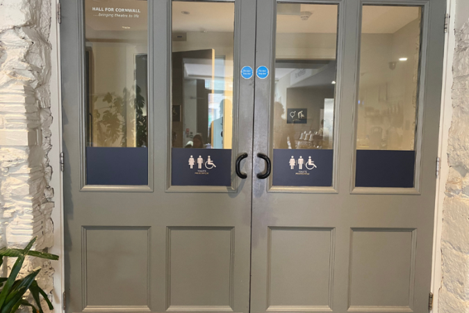 A photograph of 2 closed grey double doors, with the signs for toilets on them. Just behind the window in the doors you can see toilet doors. 