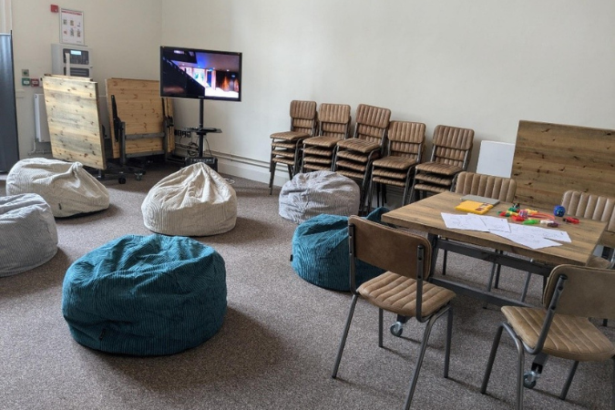 A photo of a breakout space at Hall for Cornwall, situated in a small room. Chairs have been stacked and pushed against the wall, leaving space for beanbags on the floor. There is a table and chairs in the corner, where some arts and crafts pieces and sensory toys are placed. A screen can be seen towards the back. 