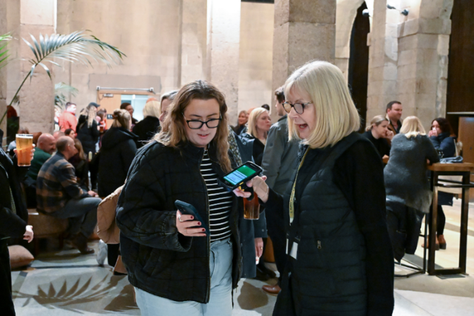 A photo of a female Usher carrying a ticket scanner, holding it up to where a customer is holding her phone up. In the background are various audience members crowded and queueing in a bar space. 