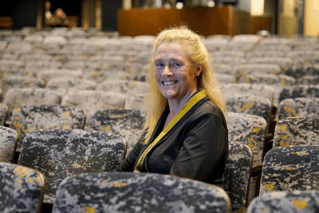 A photograph of Penny, an access usher sat in the stalls in the Cornwall Playhouse. She is wearing black and has a yellow lanyard on, smiling at the camera.