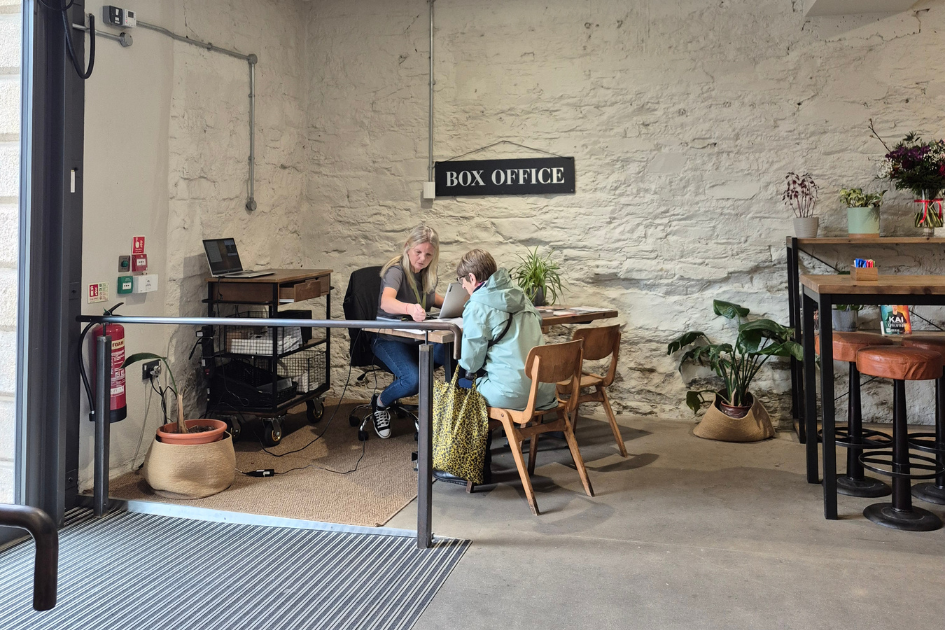 A photo of the Box Office counter, situated in the Green Room Cafe at Hall for Cornwall. To the right is a bench with stalls. To the left is the Box Office table, with a sign reading ‘BOX OFFICE’ hanging from the wall next to it. Someone is sat at the table serving a customer. 