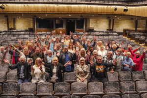 A group of HfC Volunteers stood in the stalls in the Cornwall Playhouse auditorium, they are smiling and waving at the camera.