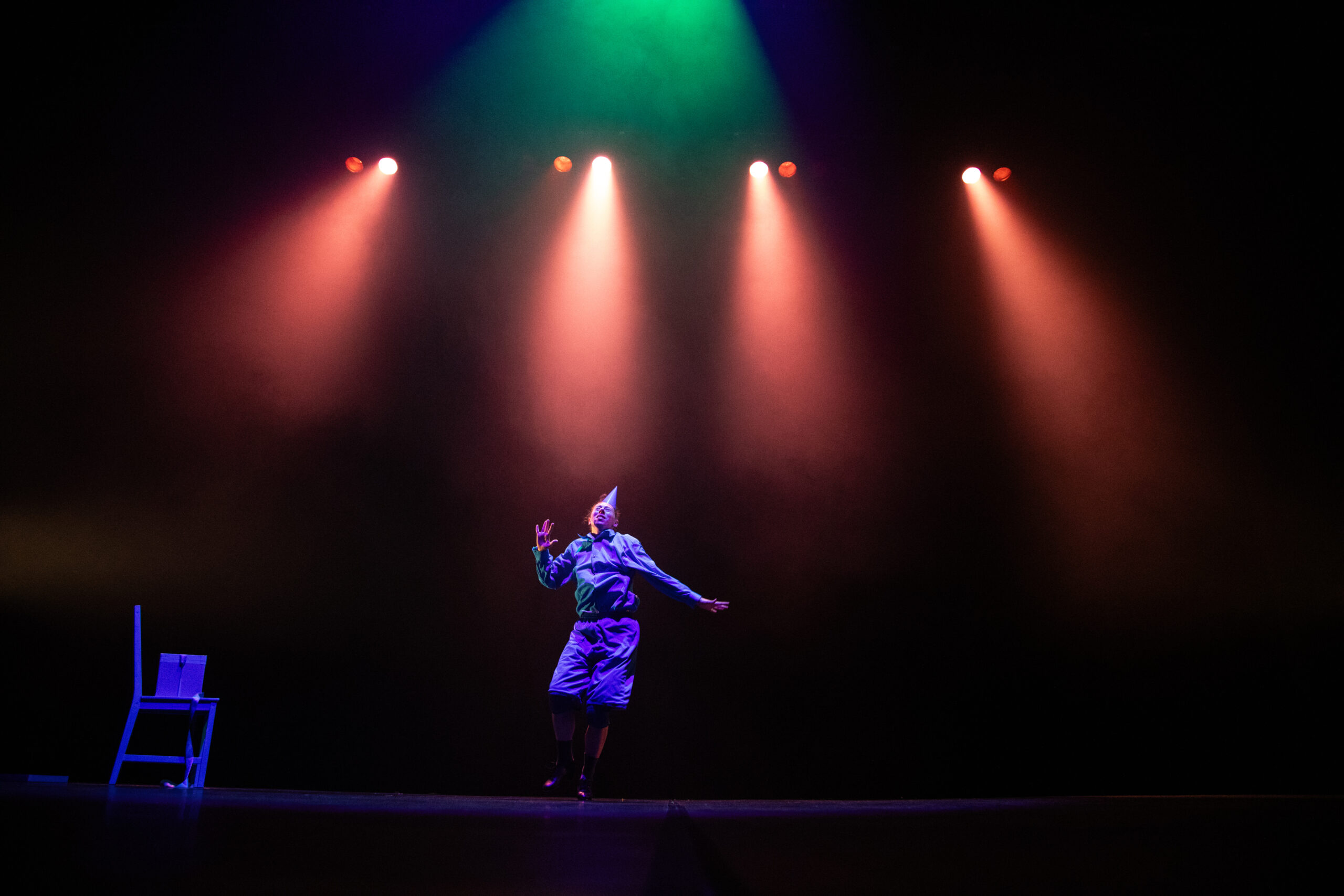 Aisha Naamani, a dancer of dual heritage and mixed ethnicity, is dancing on stage. She wears a cone shaped party hat on her head and red stage lights shine onto the stage from above.