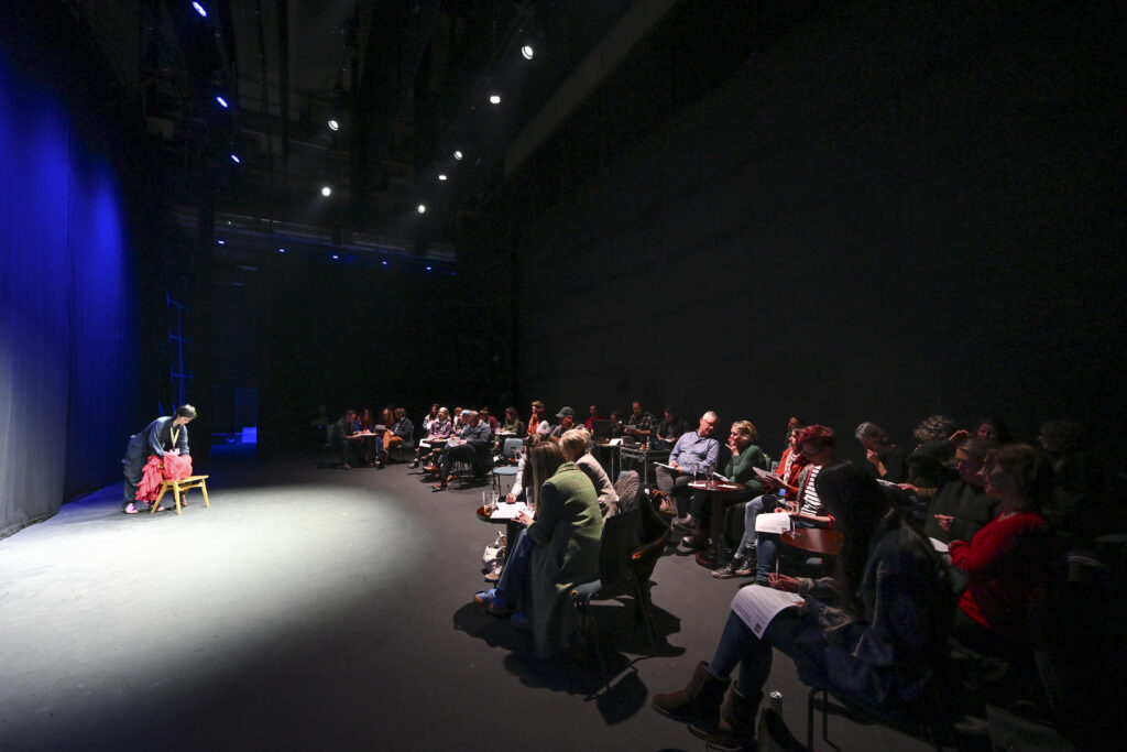A small stage bathed in light with an audience sat looking on