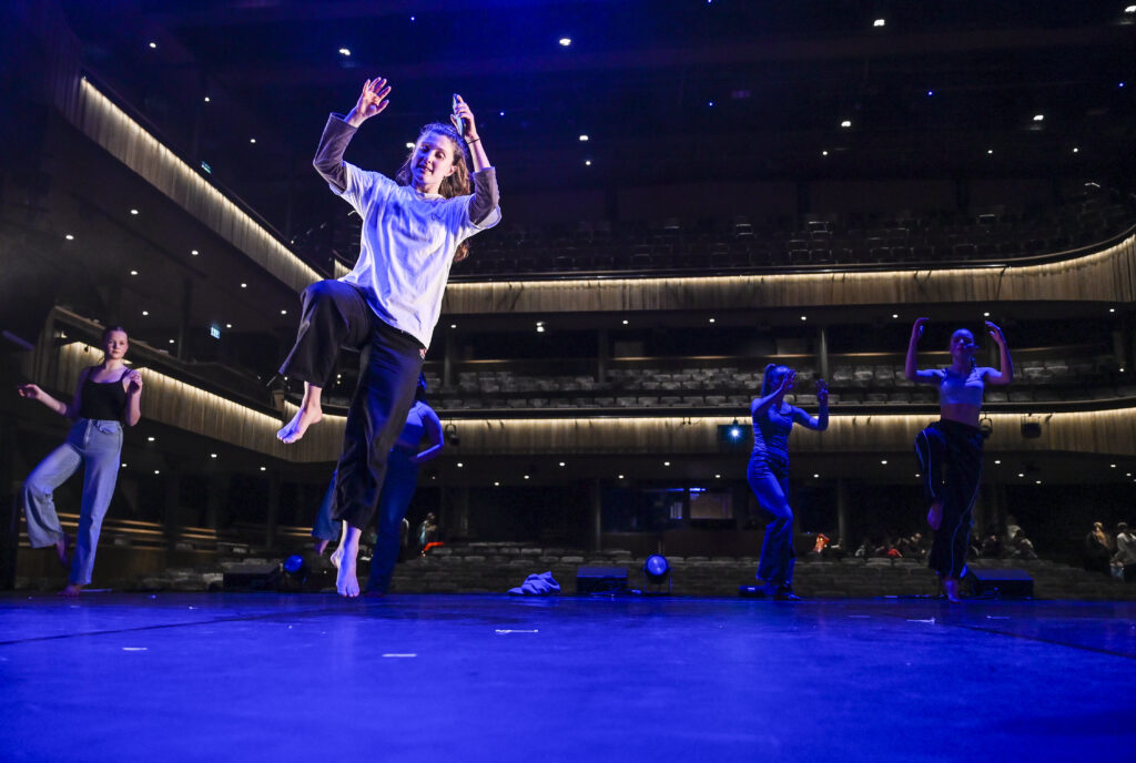 Dance artist Katie Lusby, dressed in a loose fitting white t shirt and black trousers, leads a group of teenage dancers in a rehearsal on the Cornwall Playhouse stage.