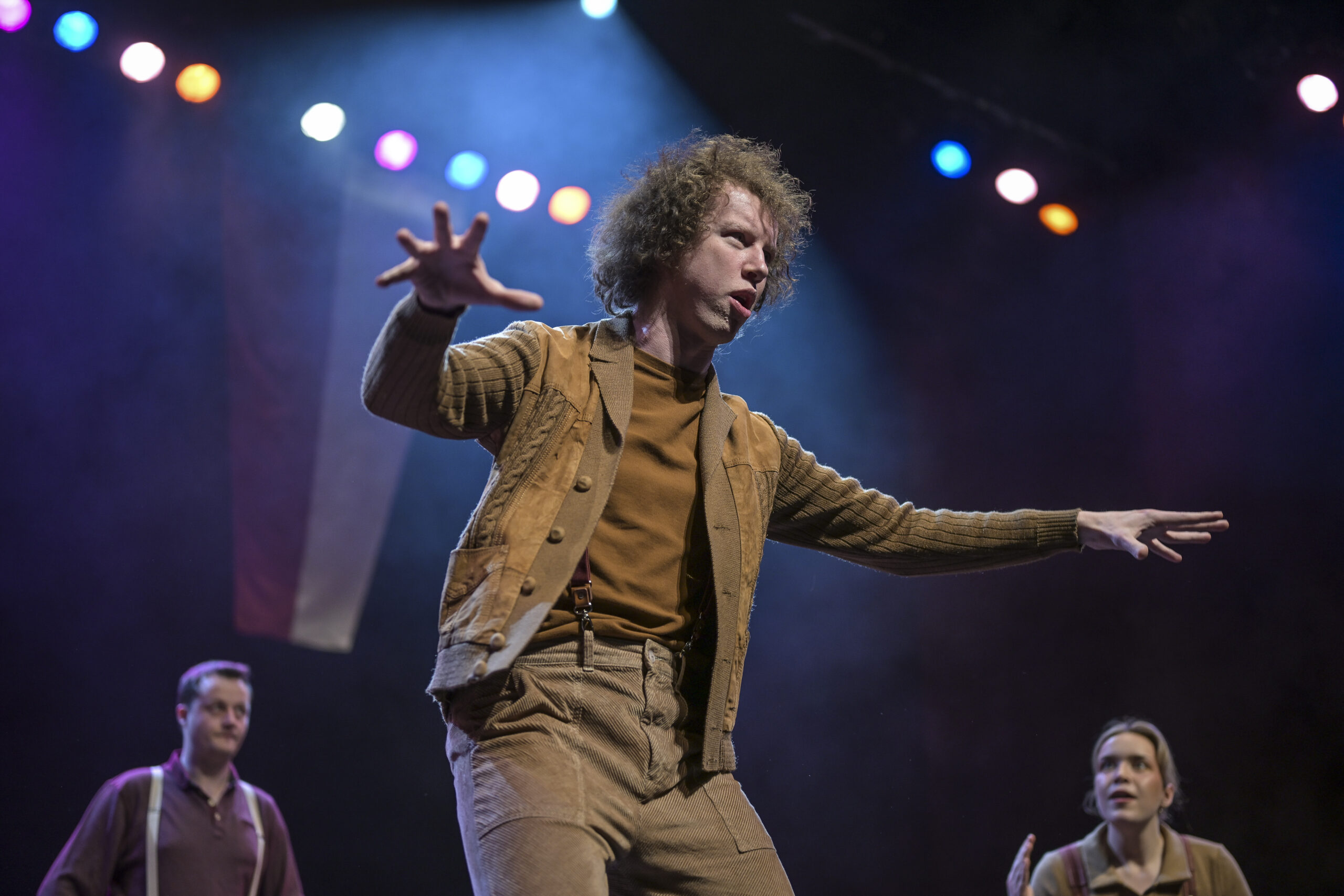 A white man with lots of curly brown hair, dressed in tan and beige clothes stands with arms outstretched to his sides. Stage lights glow behind him. 