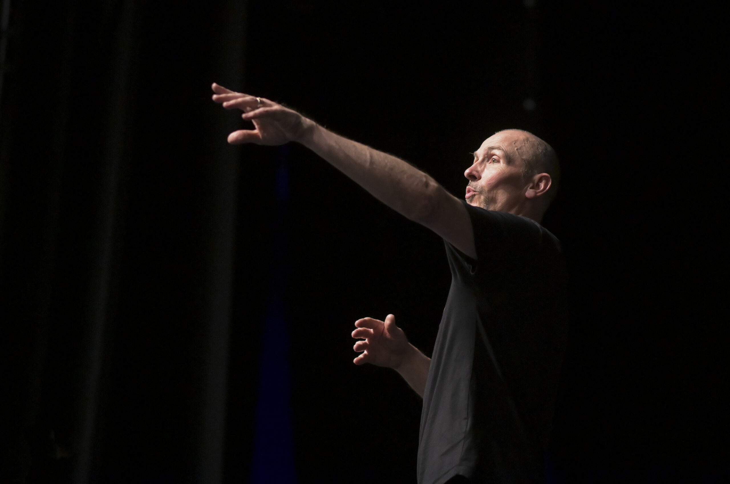 A white man with shaved head in a black T-Shirt points his arms, giving directions to the cast of Romeo and Juliet
