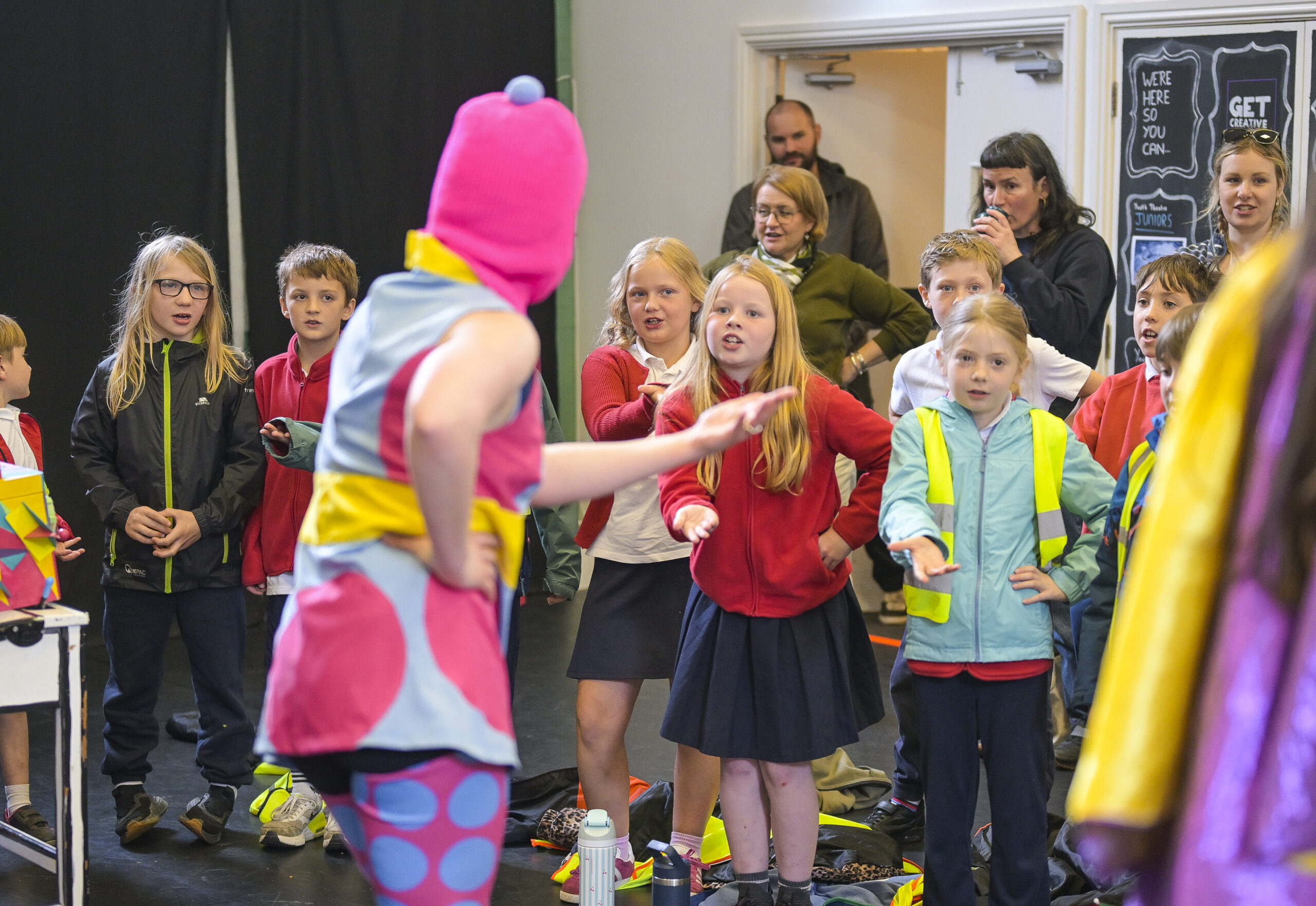 A performer from English Touring Opera dressed in brightly coloured clothes encourages the young audience to participate - a group of school children point as though telling someone off