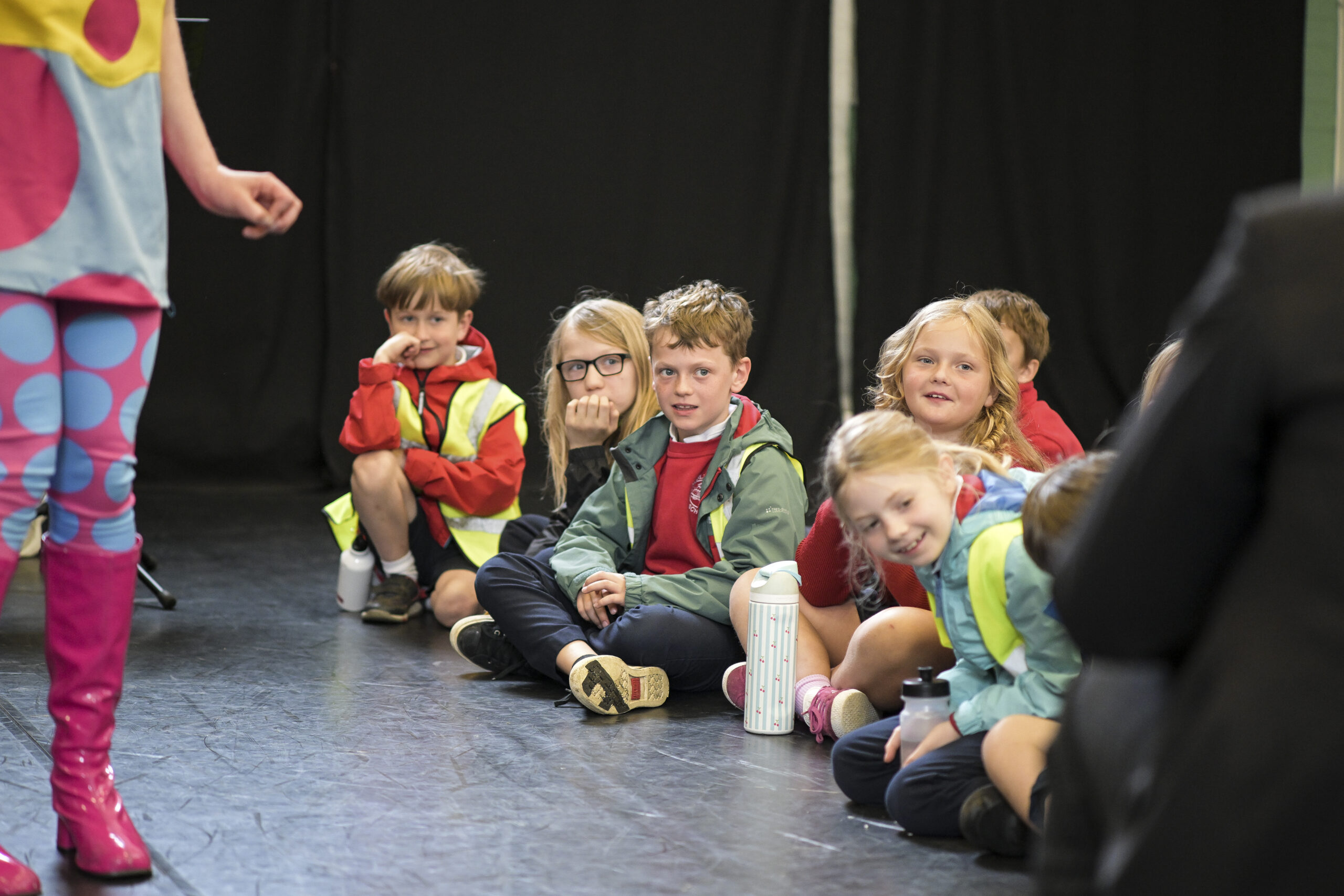 A group of school children sit cross legged on the floor - they're looking down the line smiling at each other