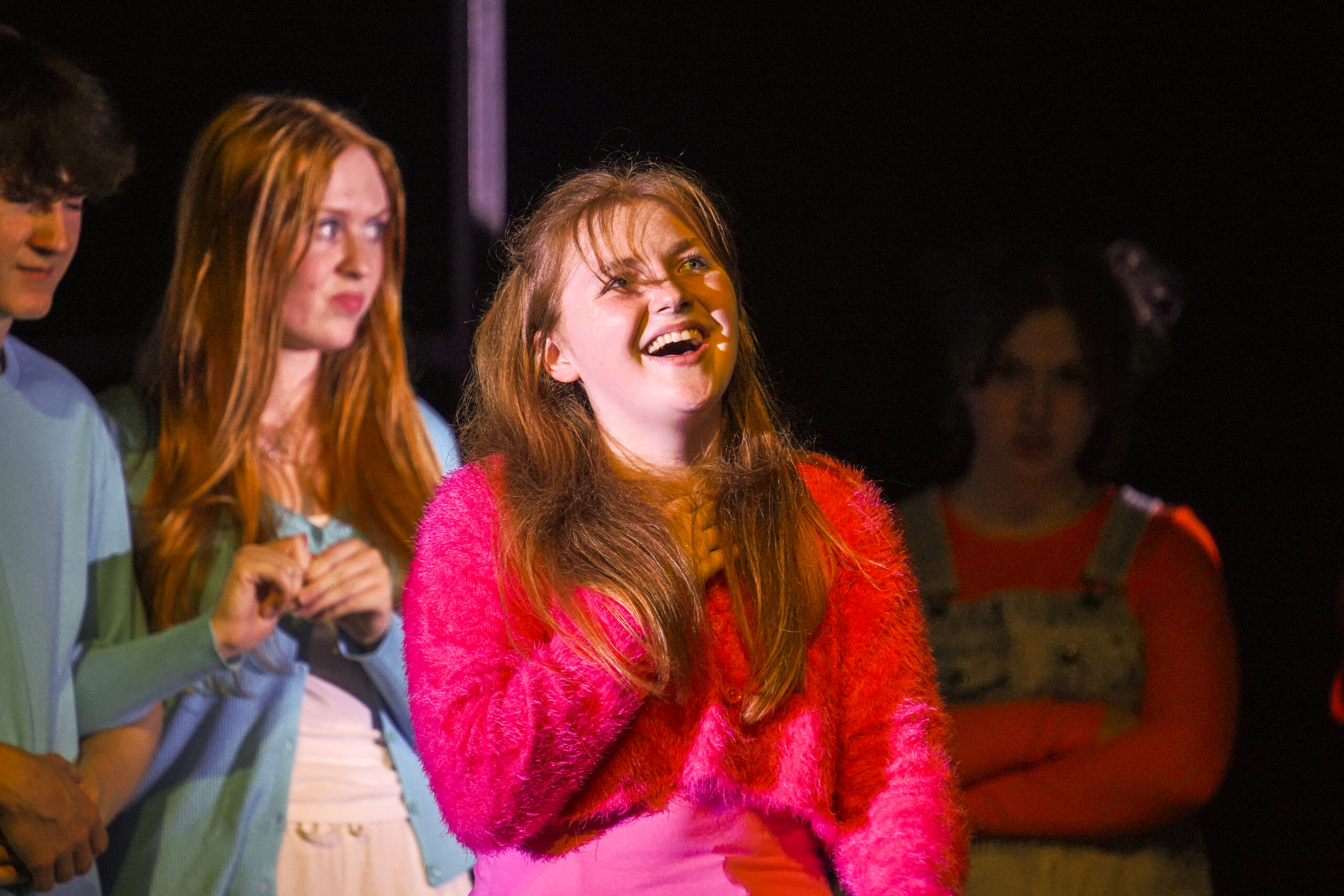 A girl with long red hair and wearing a pink jumper holds her hand to her chest with mouth open as if singing