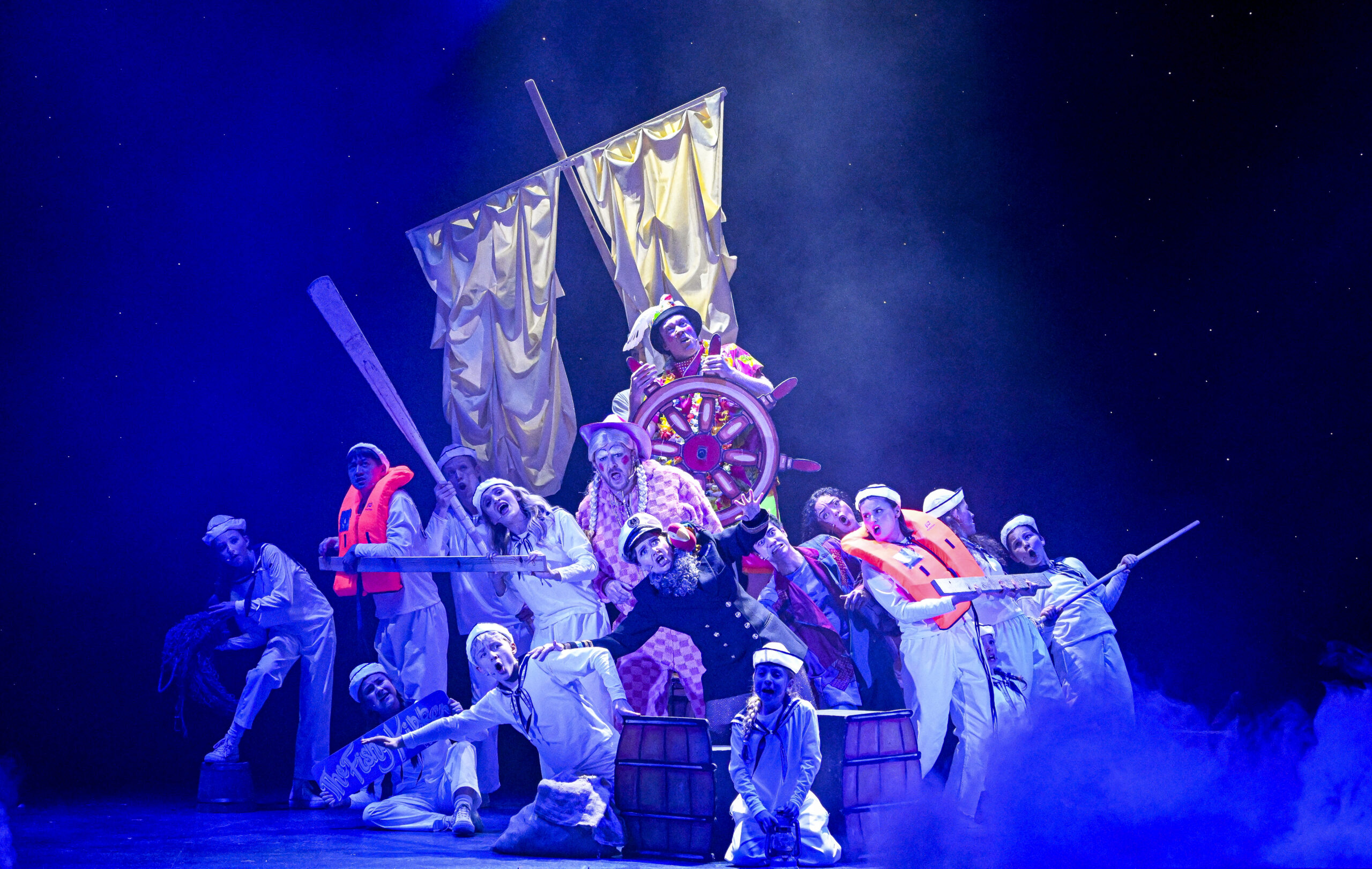 Under a blue wash of stage lights, an ensemble of actors form a group around a ship's sail and wheel. They lean to the side, singing, as if moving with the motion of the boat.