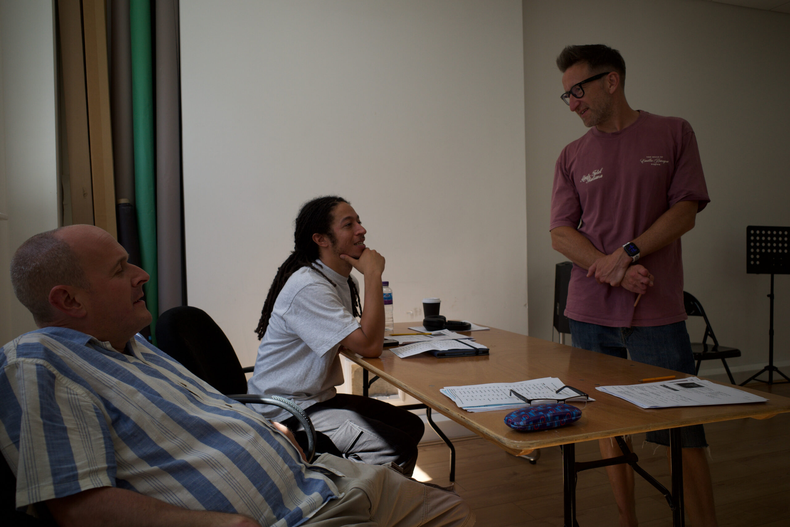 Three men, one white with a striped shirt, one black with long dreadlocked hair, one white with glasses and short brown hair all gather around a table talking