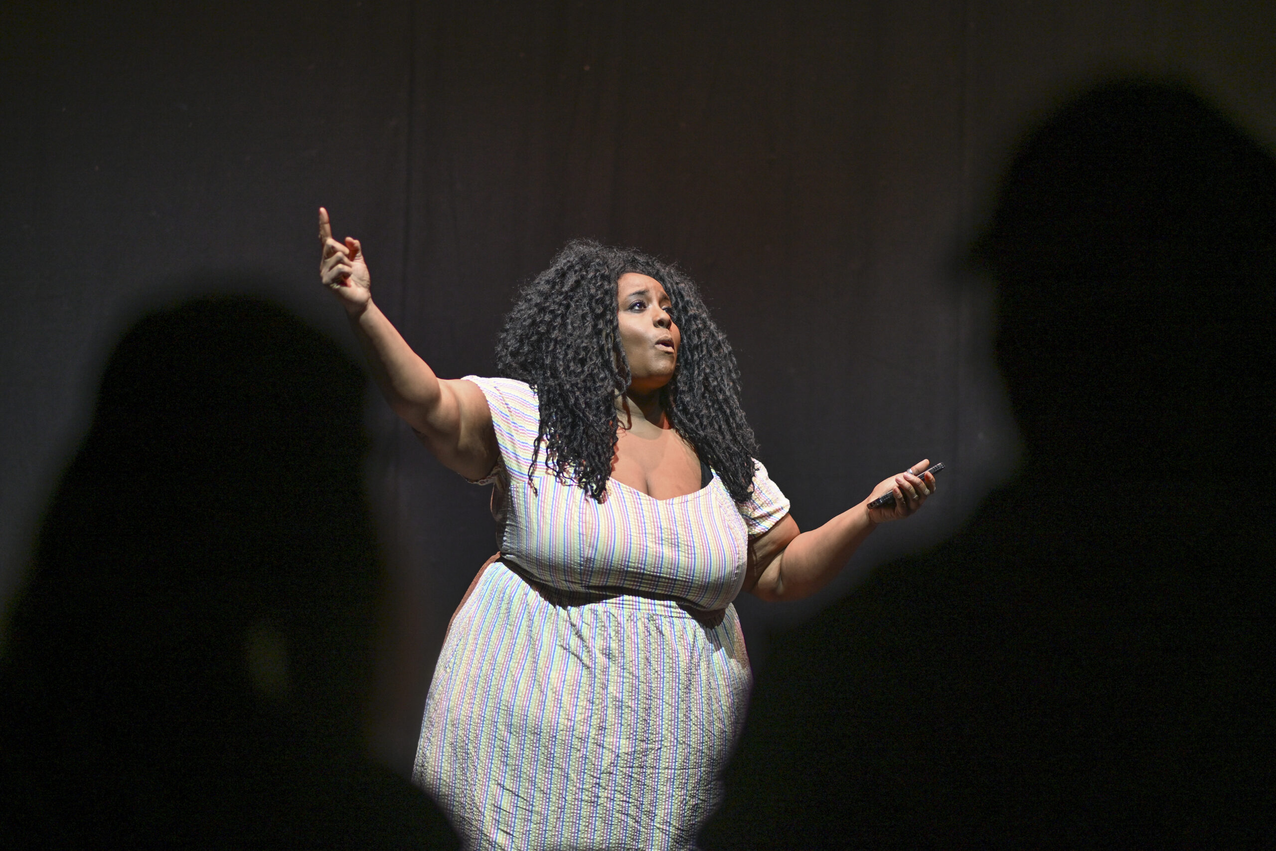 A black woman in her 40s with big, black curly hair, dressed in a white dress, holds her arms out mid-performance 