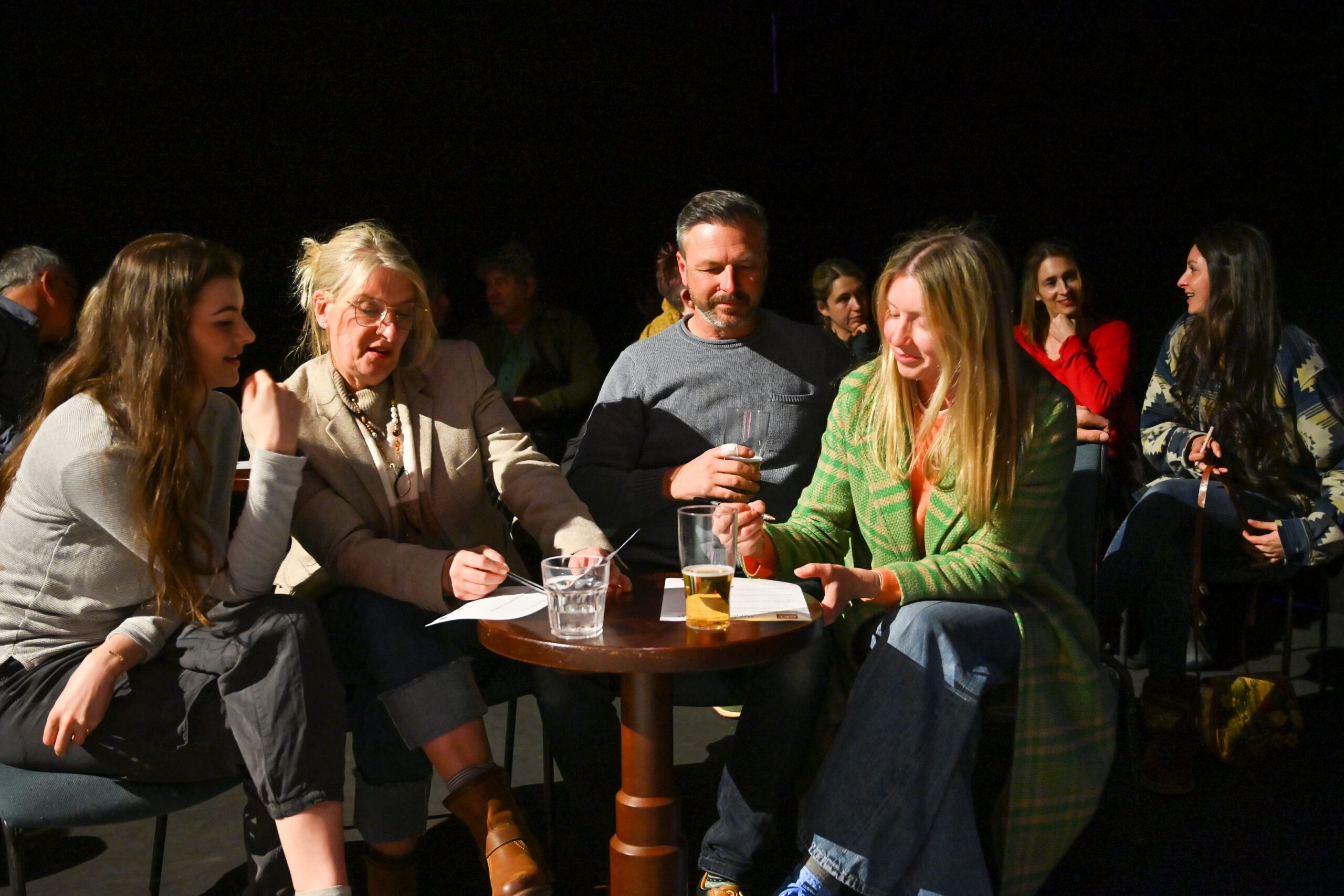 A group of people, mixed in ages and genders, gather seated around a table. They are holding paper and pens and discussing things amongst the group. Assorted drinks are sat on the table too. 