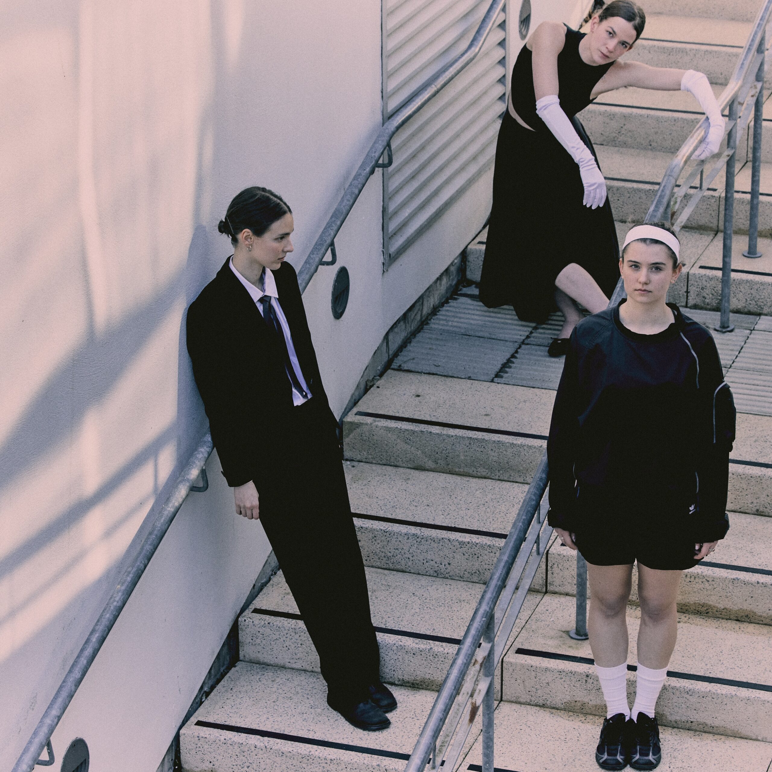 Three dancers stand on a set of concrete steps, all dressed in black and white. One, with long white evening gloves on, drapes slightly over the handrail with arms curved
